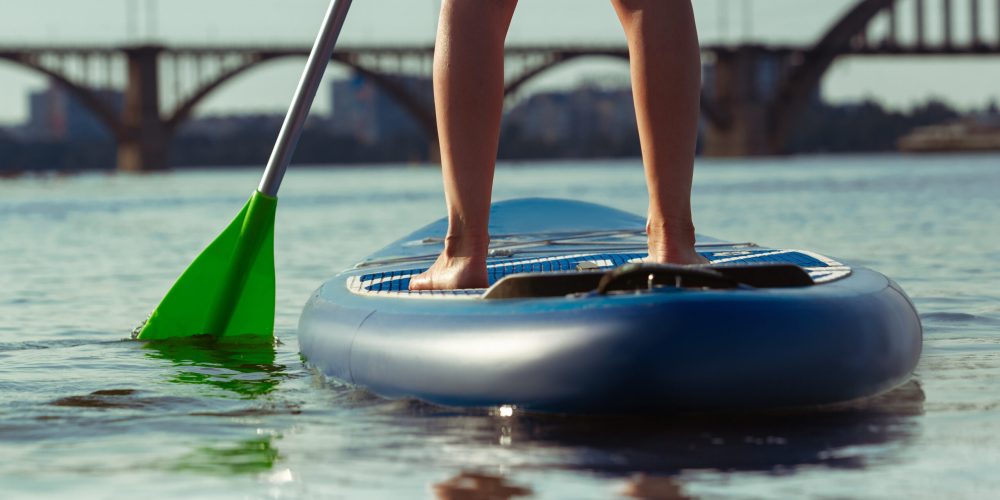 Close up legs. Young attractive woman standing on paddle board, SUP. Active life, sport, leisure activity concept. Caucasian woman on travel board in summers evening time. Vacation, resort, enjoyment.